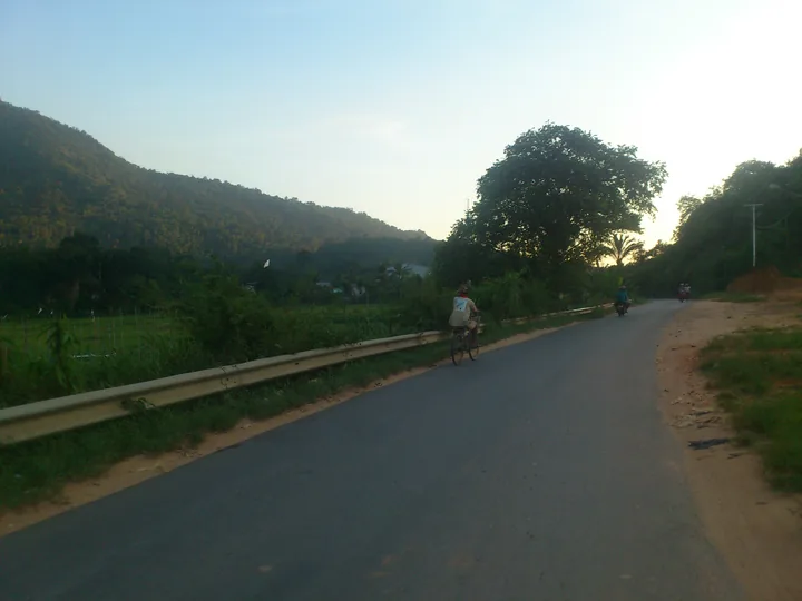 Jalan beraspal di Singkawang dengan pagar pengaman, pesepeda, dan sepeda motor.